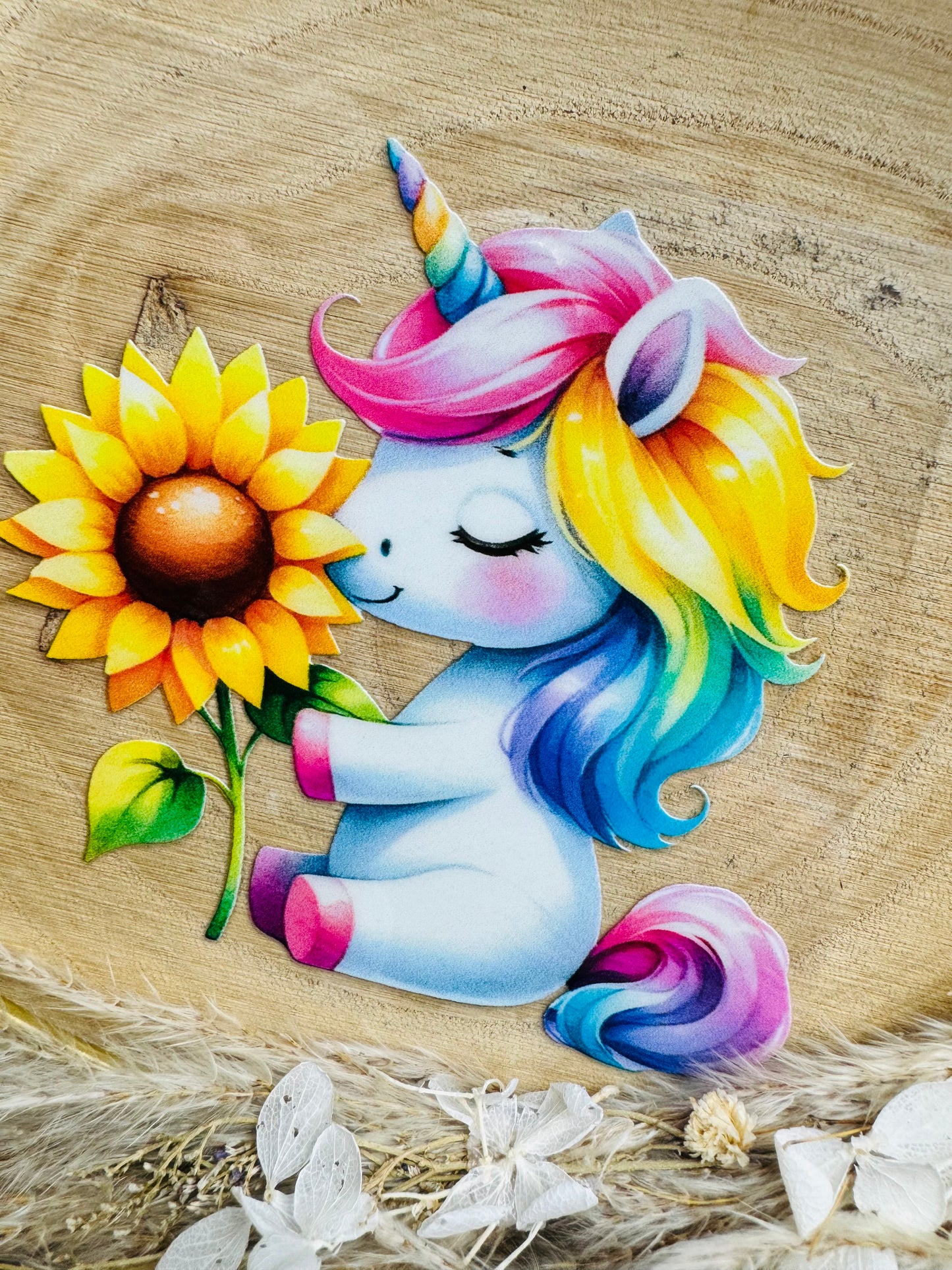 UV-Sticker mit einem kleinen sitzenden hellblauen Einhorn das an einer Sonnenblume schnuppert