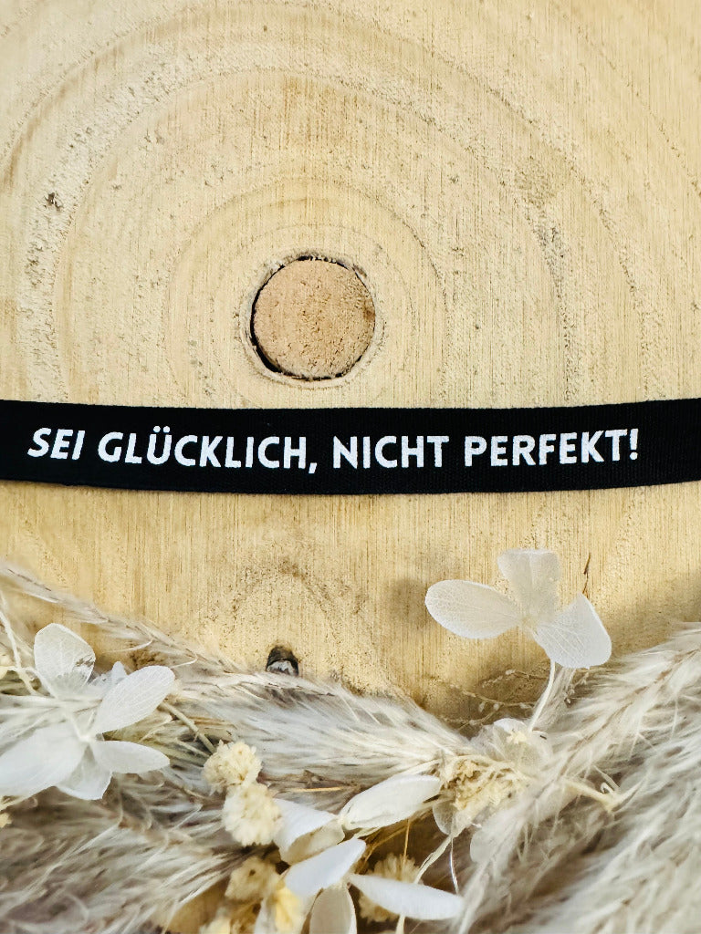 schwarze Flachkordel mit dem Spruch Sei Glücklich Komma Nicht perfekt Ausrufezeichen in Schwarz mit weisser Schrift, Breite 12 Millimeter