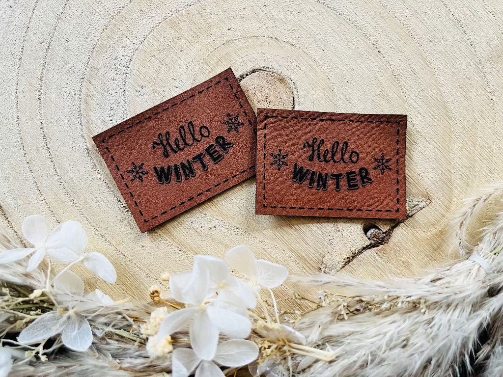 braunes Kunstleder Label mit dem Spruch Hello Winter und zwei Eiskristallen links und rechts