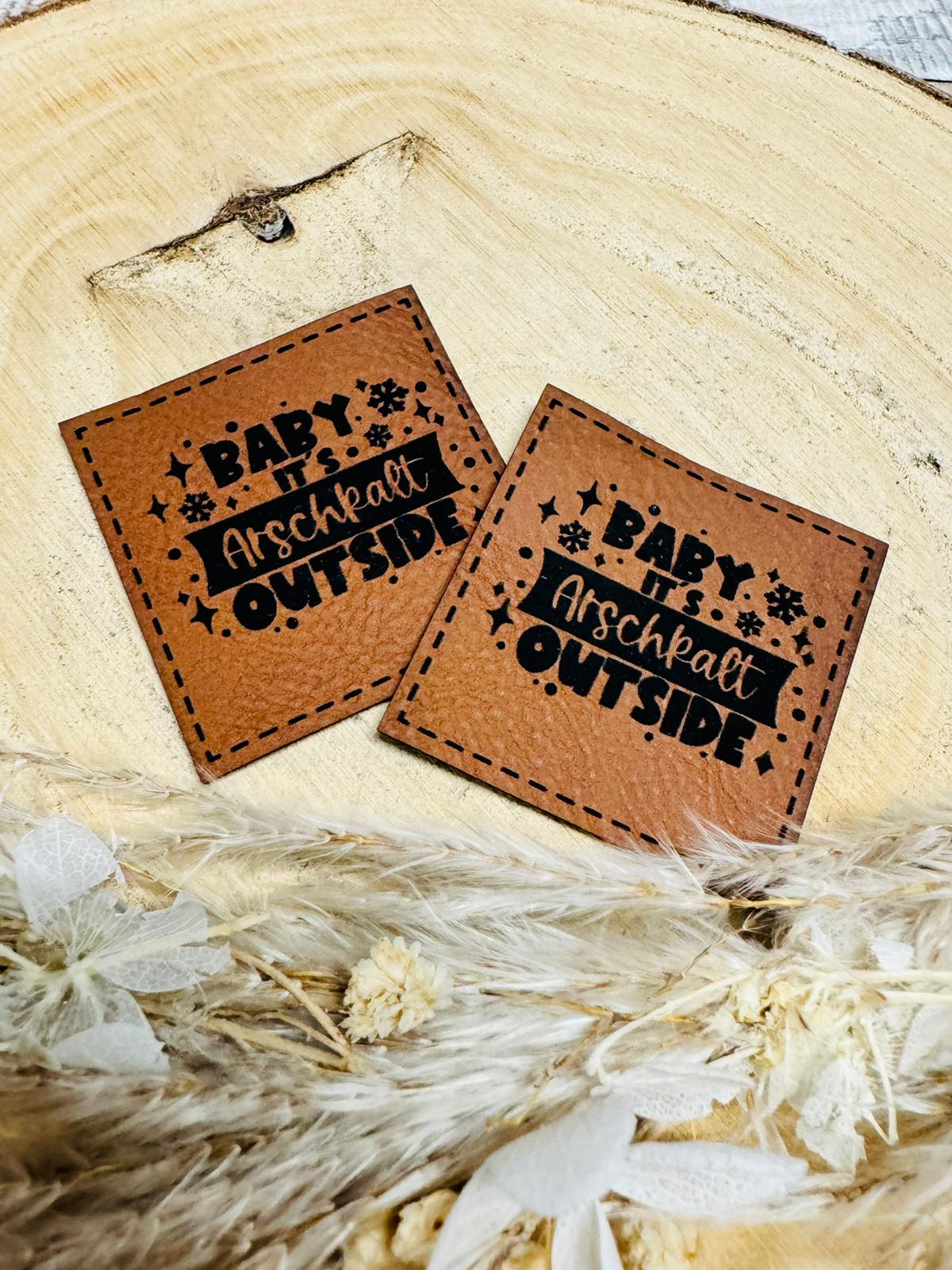braunes Kunstlederlabel mit dem Spruch Baby its Arschkalt Outside