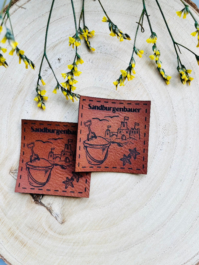 braunes PU-Label mit dem Spruch Sandburgenbauer nd einer abgebildeten Sandburg mit Eimerchen