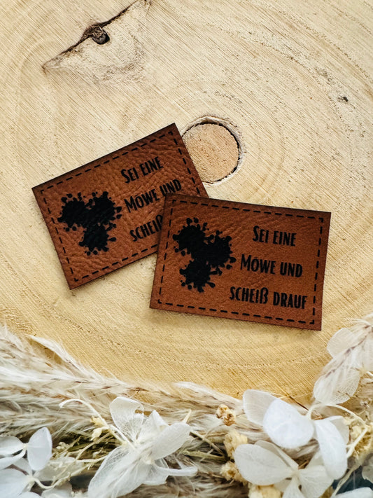 braunes Kunstlederlabel mit dem Spruch Sei eine Möe und scheiß drauf und ...Klecksen