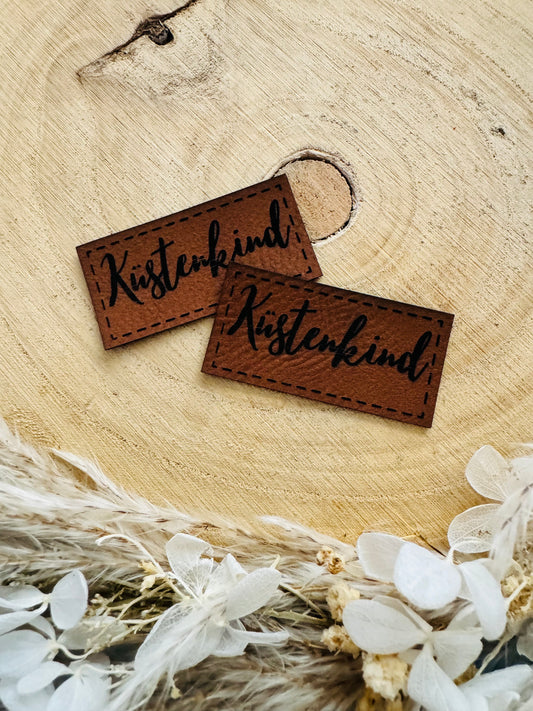 braunes Kunstlederlabel mit dem Wort Küstenkind in schwarzer Schrift