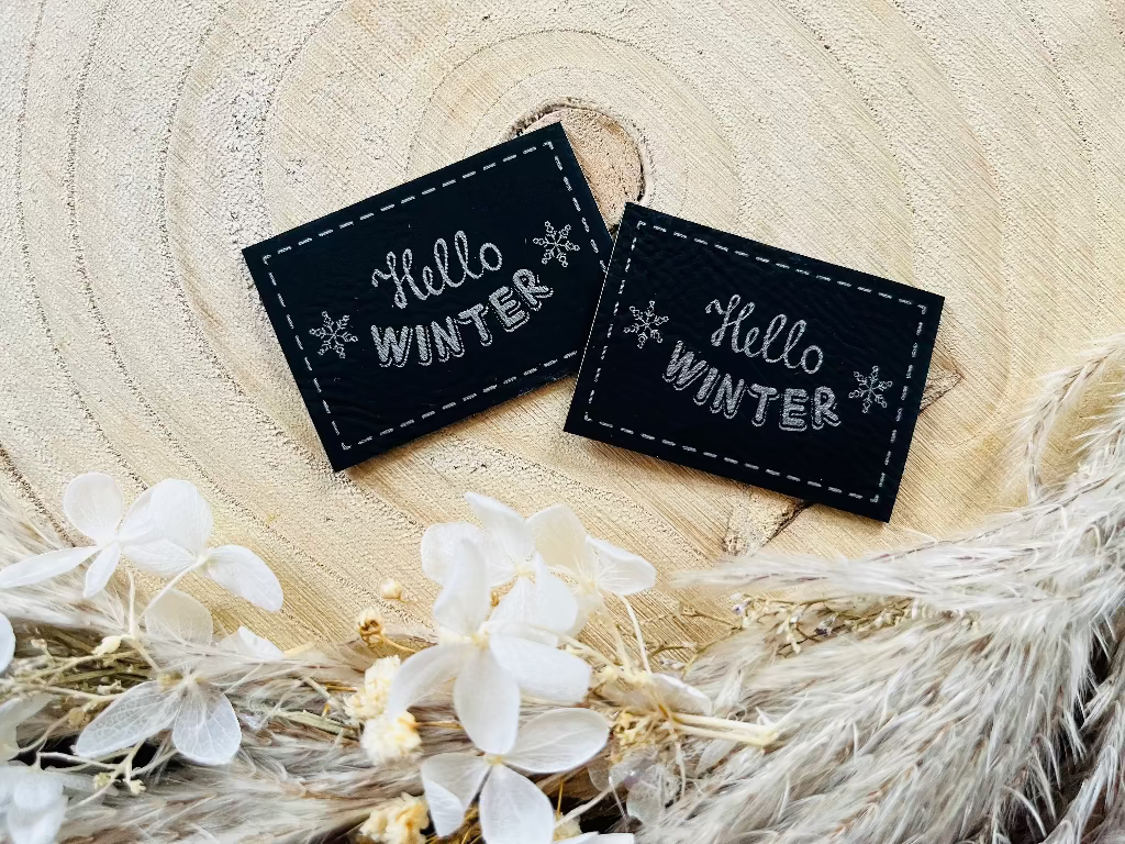 schwarzes Kunstleder Label mit dem Spruch Hello Winter und zwei Eiskristallen links und rechts in silberner Schrift