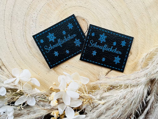 schwarzes Kunstleder Label mit dm Wort Schneeflöckchen und Eiskristallen in blau