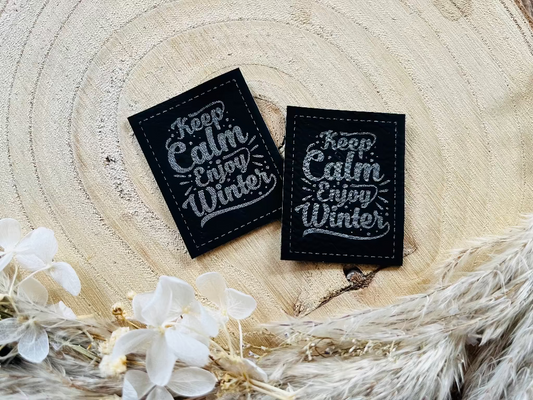 schwarzes Kunstlederlabel mit dem Spruch Keep Calm Enjoy Winter in silberner Schrift