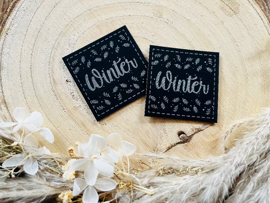 schwarzes Kunstlederlabel mit dem Wort Winter und herabgefallenen Blättern in Silber