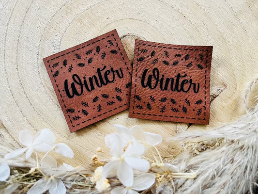 braunes Kunstlederlabel mit dem Wort Winter und herabfallenden Blättern