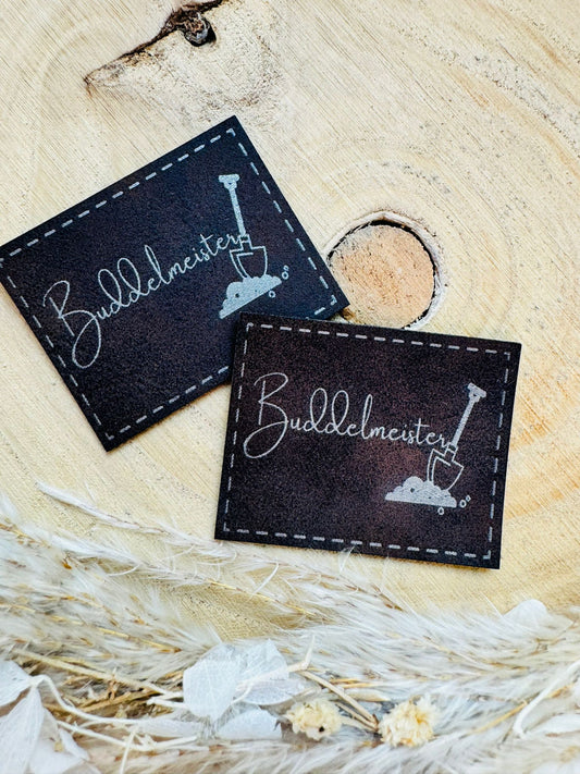 braunes PU-Label mit dem Spruch Buddelmeister und einer abgebildeten Schaufel im Sand
