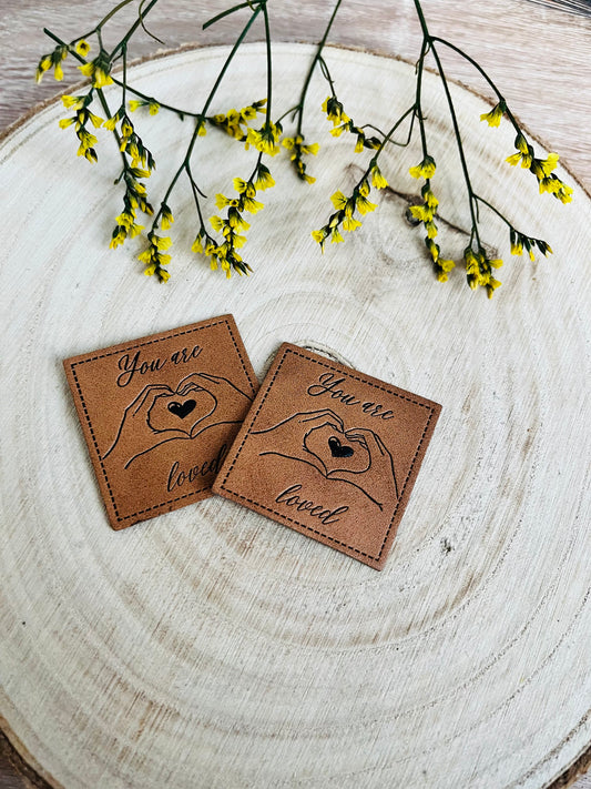 braunes kunstlederlabel mit dem spruch you are loved und zwei händen zum herz geformt