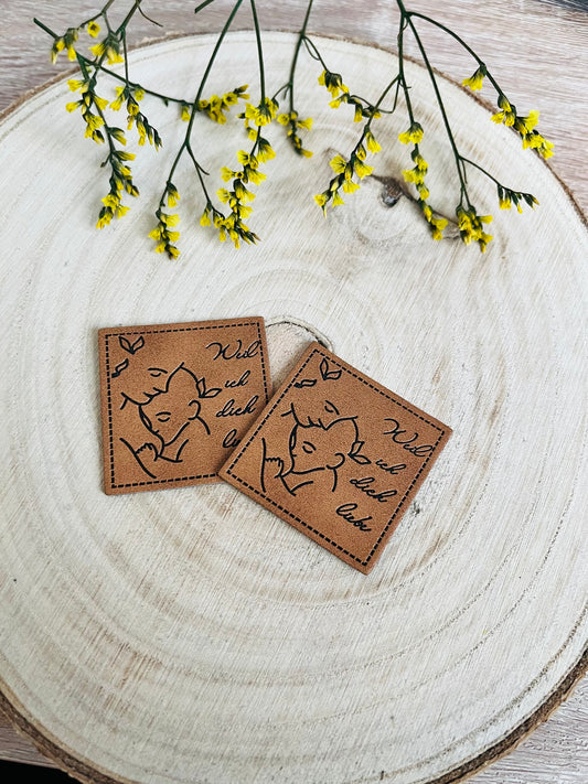 braunes kunstlederlabel mit dem spruch weil ich dich liebe und einem kleinen baby an die mutter angeschmiegt