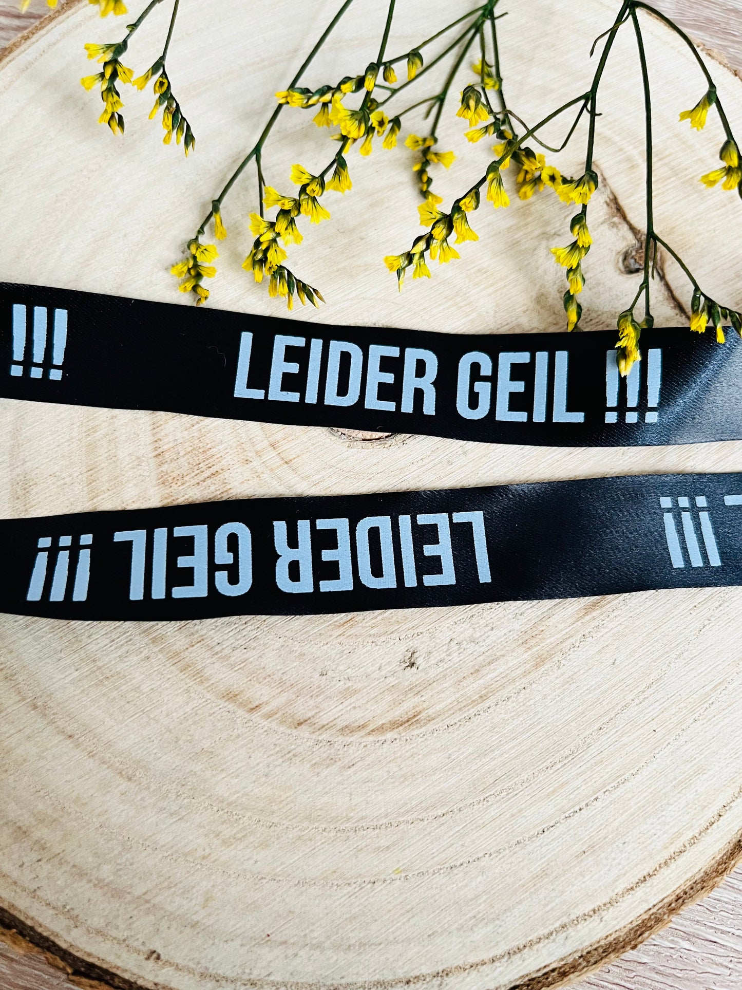 schwarzes Satinband mit dem fortlaufenden Spruch LEIDER GEIL und drei Ausrufezeichen in weisser Schrift, Breite zwei Zentimeter.