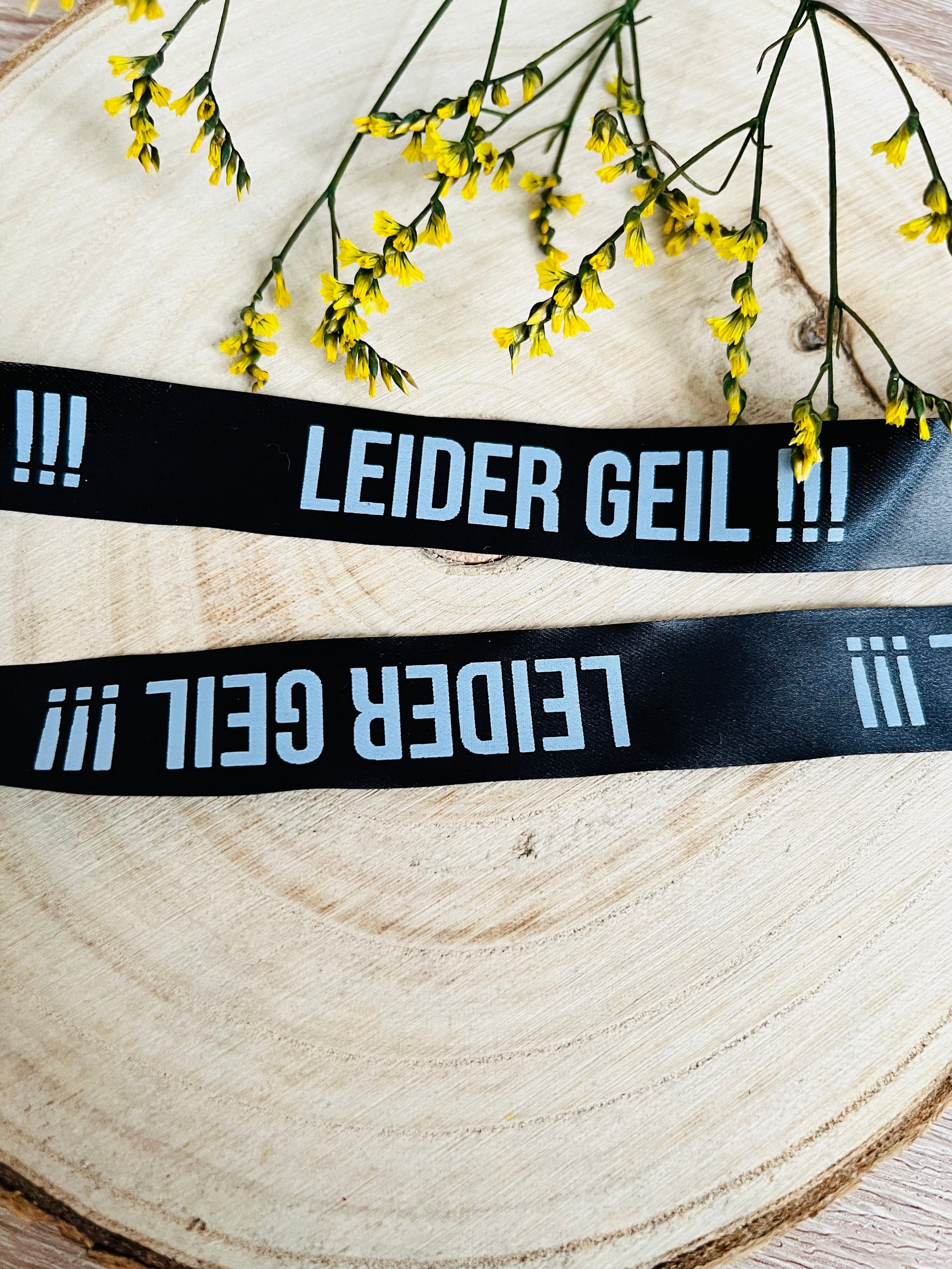 schwarzes Satinband mit dem fortlaufenden Spruch LEIDER GEIL und drei Ausrufezeichen in weisser Schrift, Breite zwei Zentimeter.