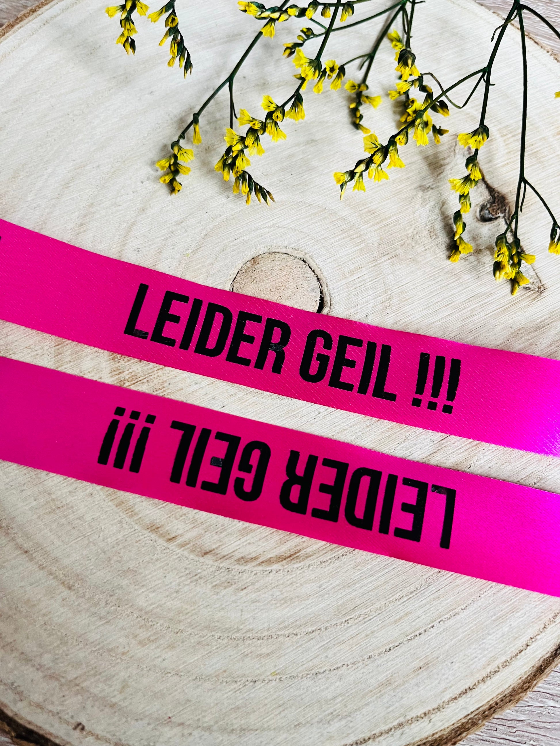 pinkfarbenes Satinband mit dem fortlaufenden Spruch LEIDER GEIL und drei Ausrufezeichen, Breite zwei Zentimeter.