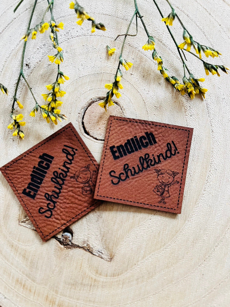 braunes Kunstlederlabel mit dem Spruch Endlich Schulkind und einem kleinen Mädchen mit Schulranzen