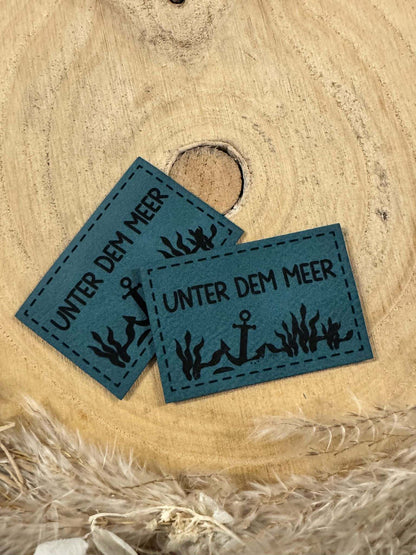 exclusives superweiches Kunstlederlabel in Türkis mit dem Spruch Unter dem Meer und einem Anker auf dem Meeresboden abgebildet