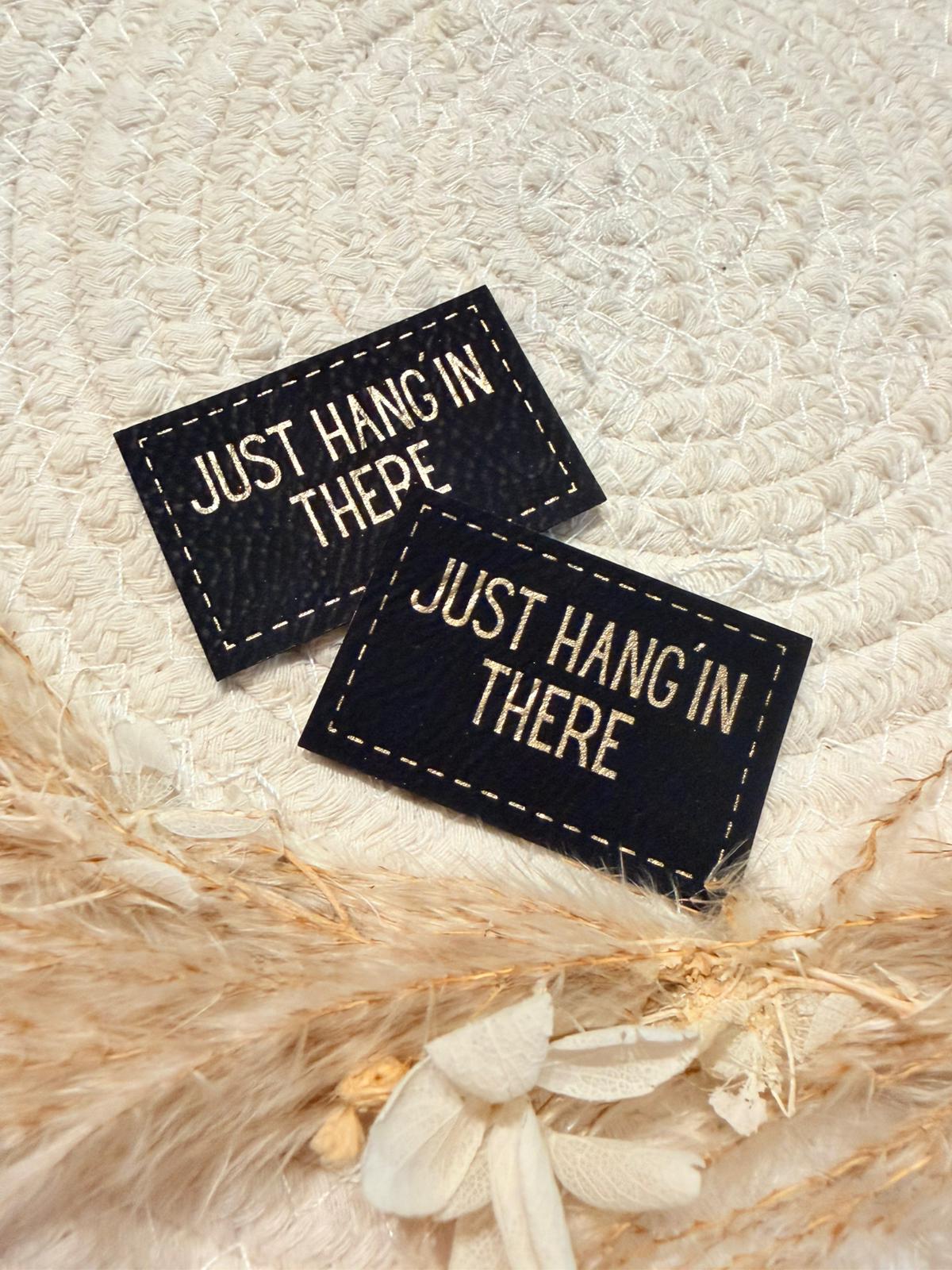 superweiches schwarzes Kunstlederlabel mit dem Spruch Just Hang In There in goldenen Buchstaben