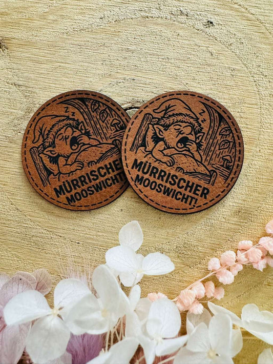 superweiches rundes braunes Kunstlederlabel mit dem Spruch mürrischer Mooswicht und einem kleinen im Bett liegenden Mooswichtel