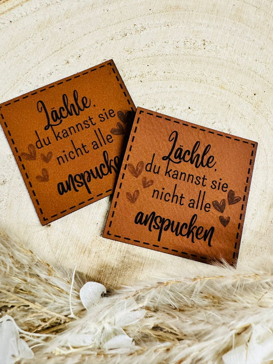 superweiches braunes Kunstlederlabel mit dem Spruch Lächle, du kannst sie nicht alle anspucken in schwarzer Schrift